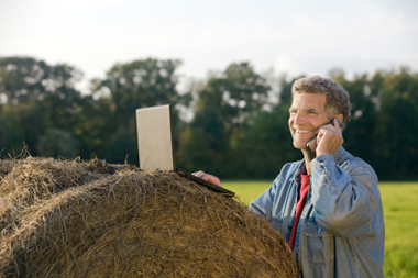 farmer in a field, speaking on a cell phone, with an open laptop on a hay bale.