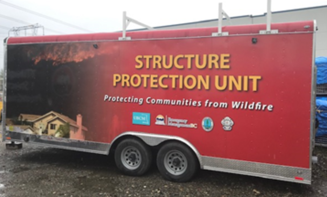 Side view of a smaller, red trailer reading "structure protection unit" and parked in a gravel lot