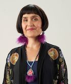 Headshot of Dr. Daniele Behn Smith who is Deputy Provincial Health Officer of Indigenous Health in British Columbia. Adult woman with dark brown bob haircut and bangs smiling slightly and wearing matching pink purple and light blue earings and necklace made of hide, seed beads, and feathers. Her jacket is black with beaded motifs.