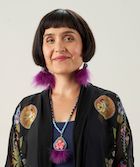 Foto da cabeça da Dra. Daniele Behn Smith, vice-diretora provincial de saúde de saúde indígena na Colúmbia Britânica.  Mulher adulta com corte de cabelo bob marrom escuro e franja sorrindo levemente e usando brincos rosa roxo e azul claro combinando e colar feito de couro, miçangas e penas.  Sua jaqueta é preta com motivos de miçangas.