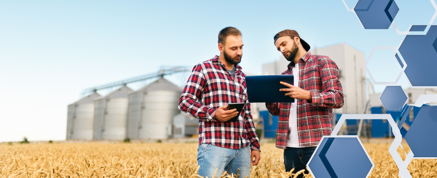 Two men in plaid shirts standing in a field while reviewing something on their tablets, with granaries in the background