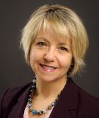 Headshot of Dr. Bonnie Henry who is BC's Provincial Health Officer. She is a white woman with blonde hair reaching her chin and bangs. She is wearing a dark coloured blazer, a pink blouse, and a necklace of round blue and brown glass beads with white dots.