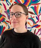 Headshot of Dr. Kate Jongbloed who is a post doc in the BC Office of the Provincial Health Officer. She is a white woman with dark hair in a low pony tail wearing black rimmed glasses and a black sweatshirt. Behind her is an otomi embroidery wall hanging with vibrant colours.