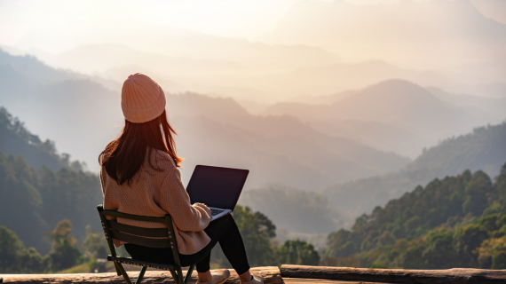 A woman sits with her laptop while looking out at the mountains 