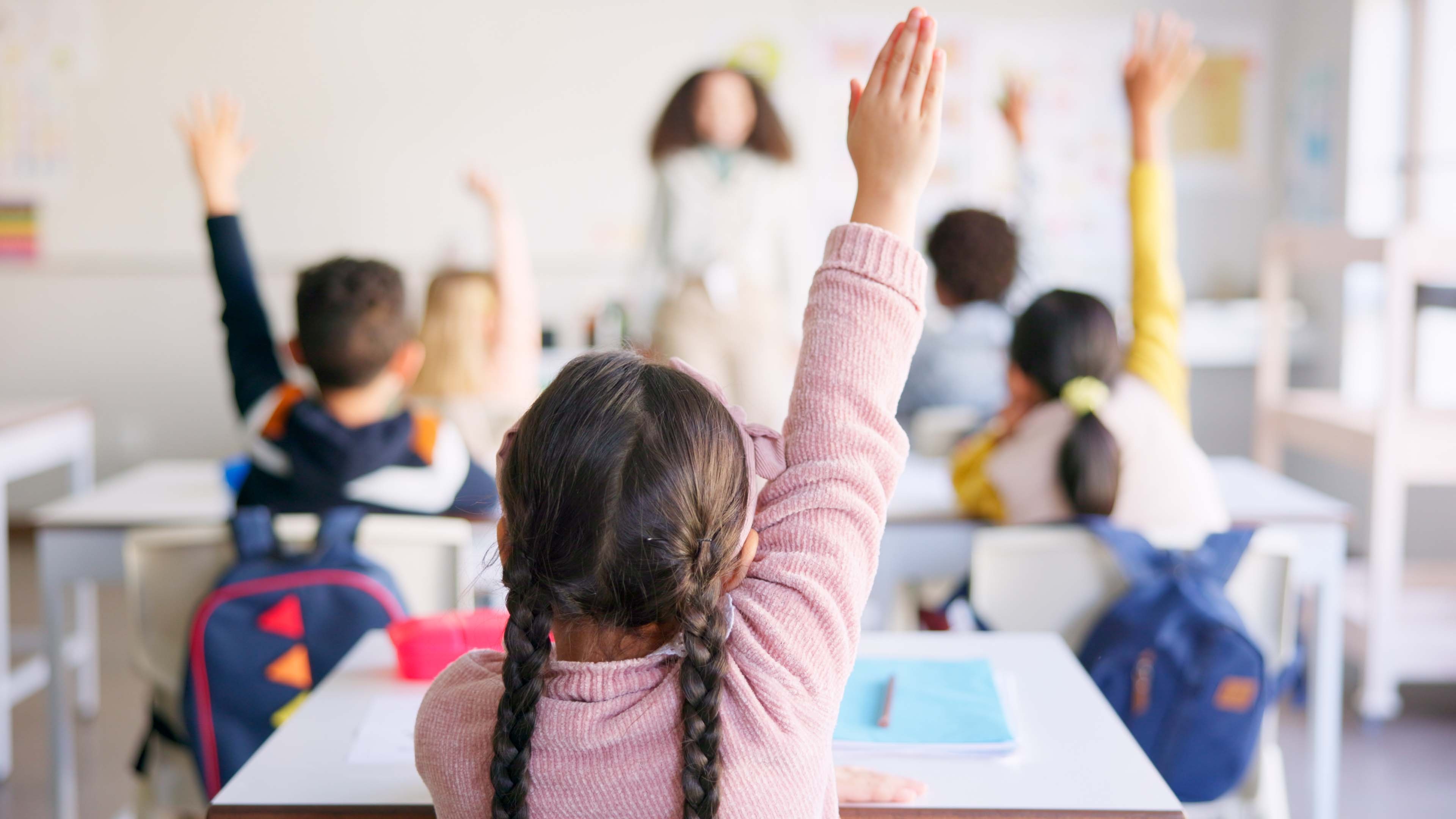 Children sitting at desks in a classroom, raising hands. 