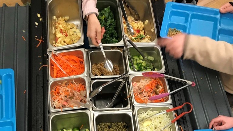 Children reaching with utensils to serve themselves at a salad bar.