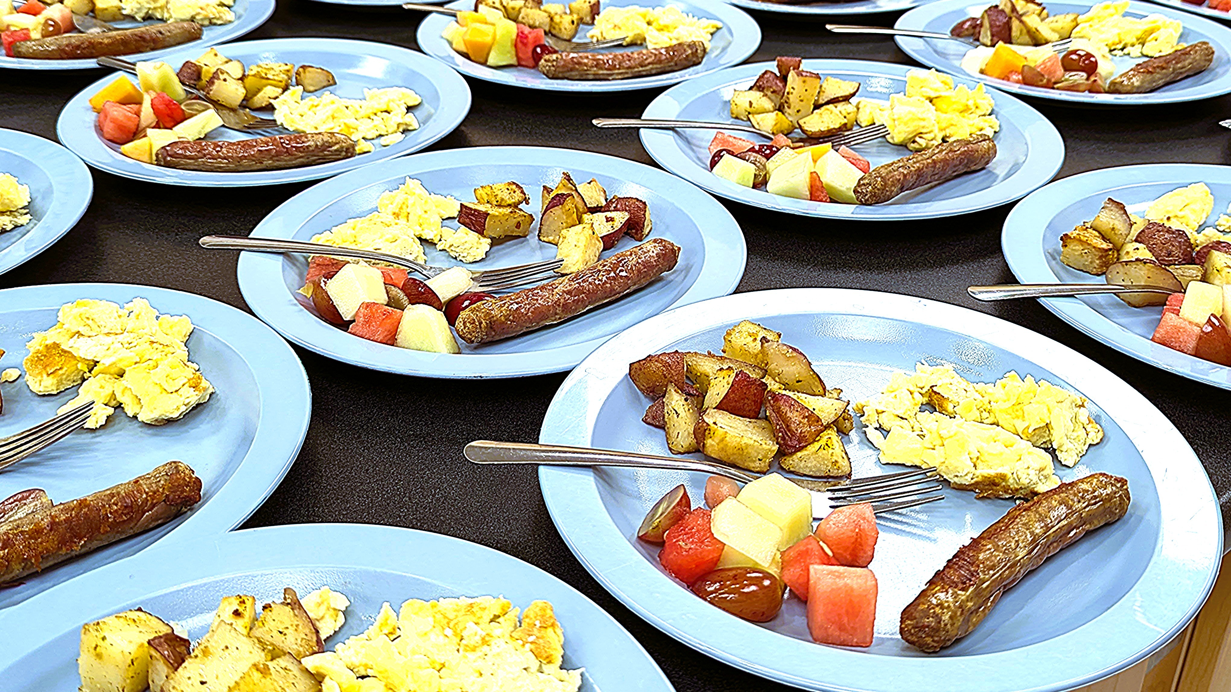 Plates of breakfast including sausage, scrambled eggs, potatoes and fruit.