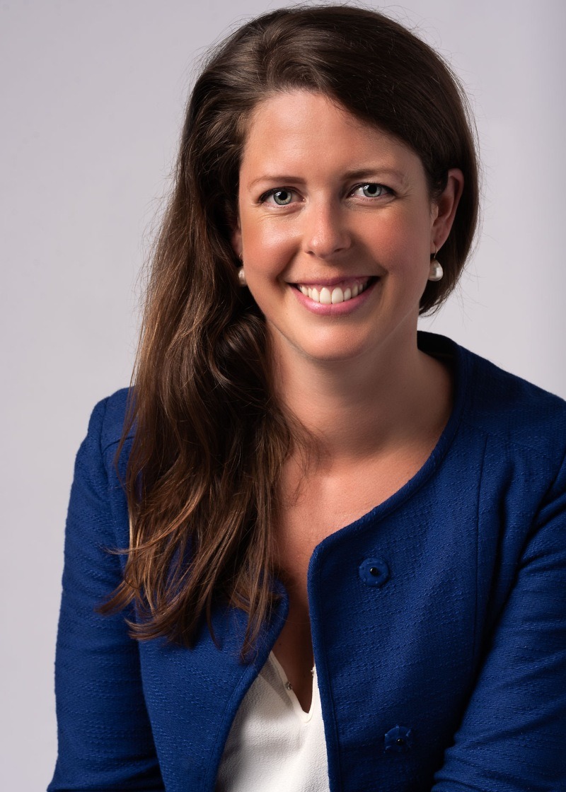 A woman with light skin tone wearing a blue blazer over a light-coloured top, photographed indoors against a plain light background. She has long brown hair parted more to one side, is smiling, and is looking directly at the camera.