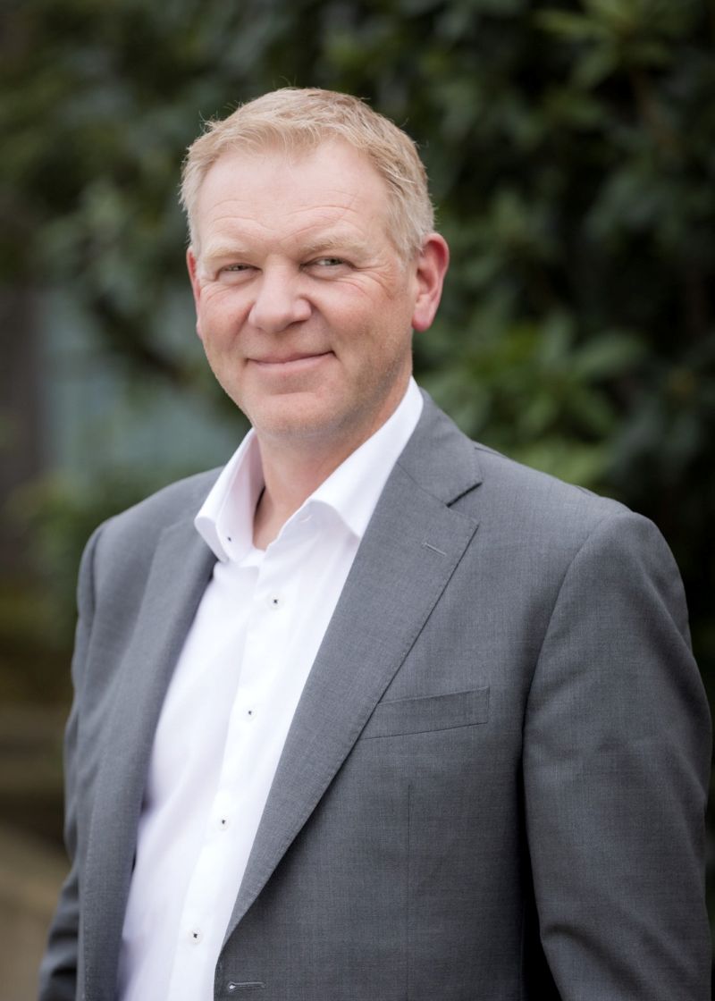 A man with light skin tone wearing a grey suit jacket over a white collared shirt, standing outdoors with greenery in the background. He has short light-coloured hair and is looking at the camera with a slight smile.