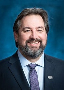 A man with light skin tone wearing a dark suit jacket, light dress shirt, and purple tie, photographed indoors against a blue studio-style background. He has short dark hair with some grey, a full beard, and is smiling while looking at the camera.