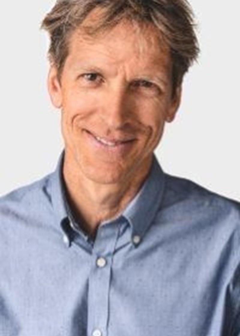 A man with light skin tone wearing a light blue button-up shirt, photographed indoors against a plain white background. He has short brown hair and is smiling while looking at the camera.