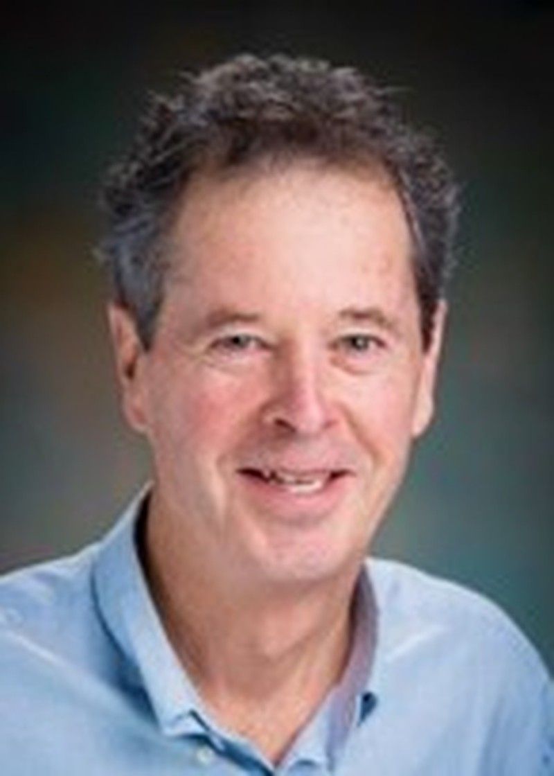 A man with light skin tone wearing a light blue collared shirt, photographed indoors against a softly blurred dark background. He has short dark hair with some grey and is smiling while looking at the camera.