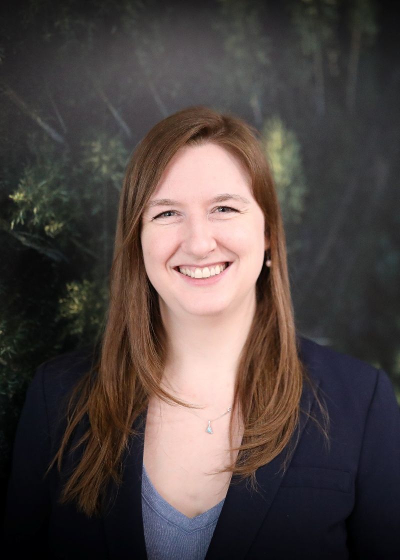 A woman with light skin tone wearing a dark blazer over a light-coloured top, standing outdoors against a background of trees and greenery. She has long light brown hair worn down and is smiling while looking at the camera.