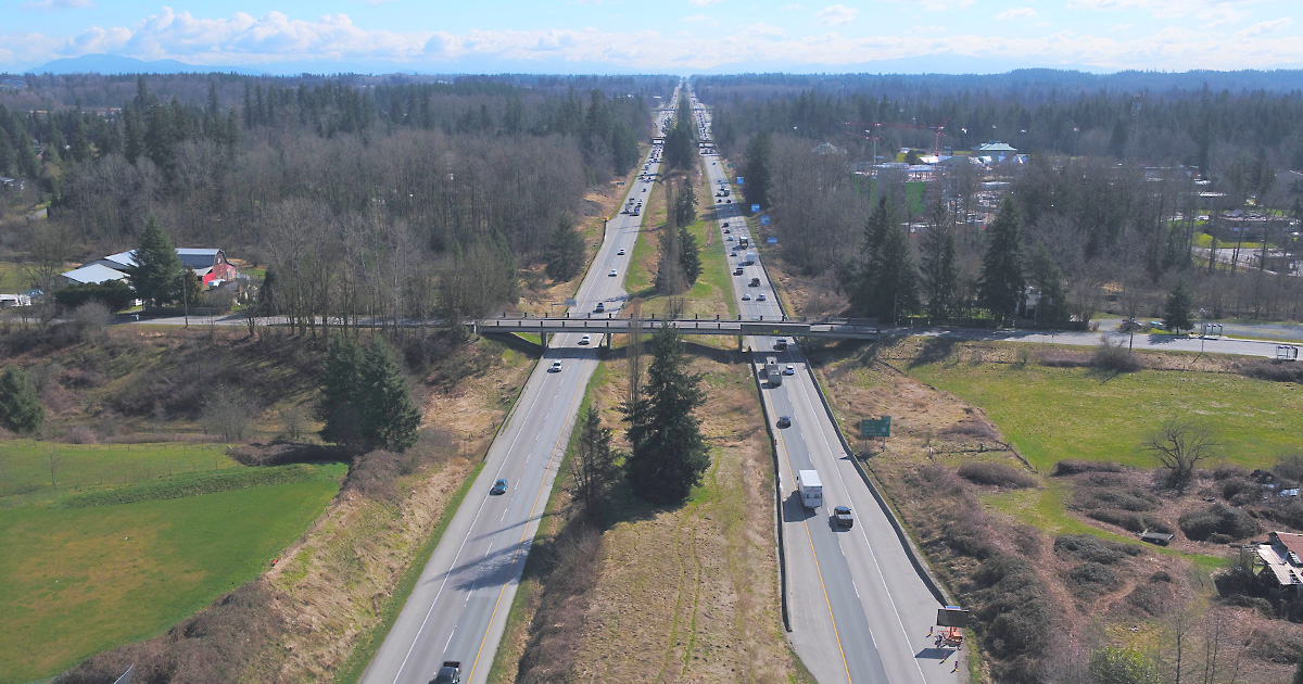 Photo of Glover Road showing two lanes each direction with an overhead bridge, located in a farmland area with coniferous and deciduous trees flanking the highway into the distance.