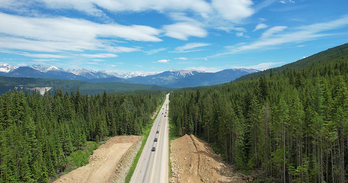 Selkirk project site showing building up of land on either side of the road to accommodate the future 4-laning and rest area. Shows the 2-lane highway with traffic flowing through the picturesque heavily evergreen forested area with snow-capped mountains reaching into blue sky with clouds on the horizon. 