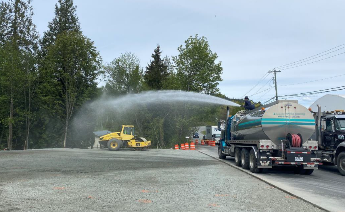 Water truck spraying water on a construction site with a compactor in the background and trees in the distance.