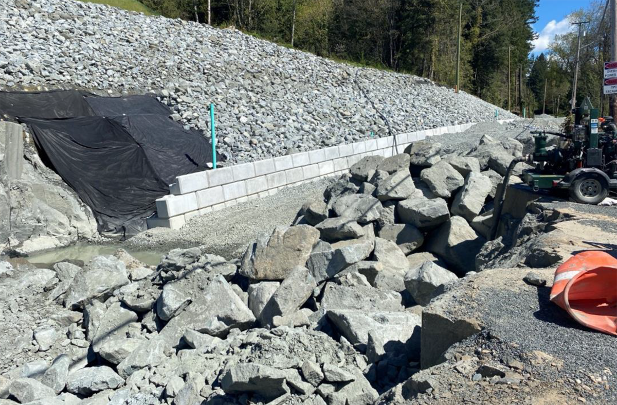  A construction site features a retaining wall made of concrete blocks, piles of large rocks, a gravel path, and an orange traffic cone. Dense forest is seen in the background.