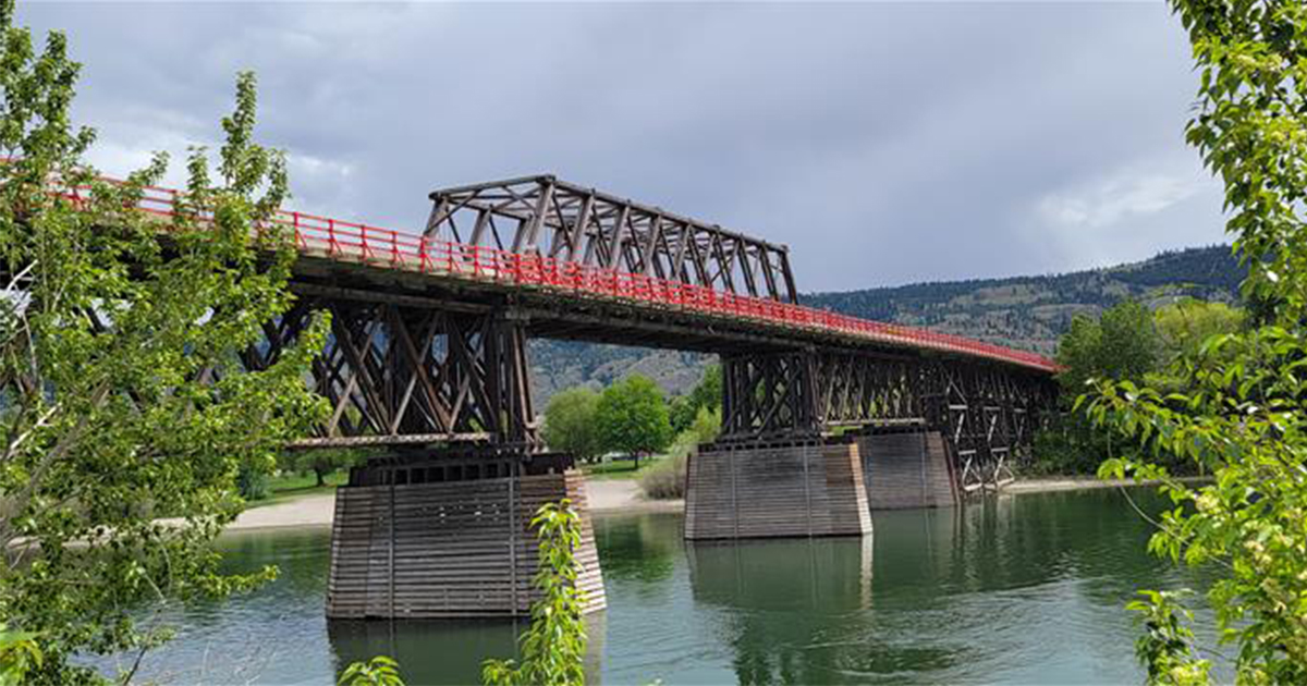 Aging red and black truss bridge crossing river with hill in the backgroud. 